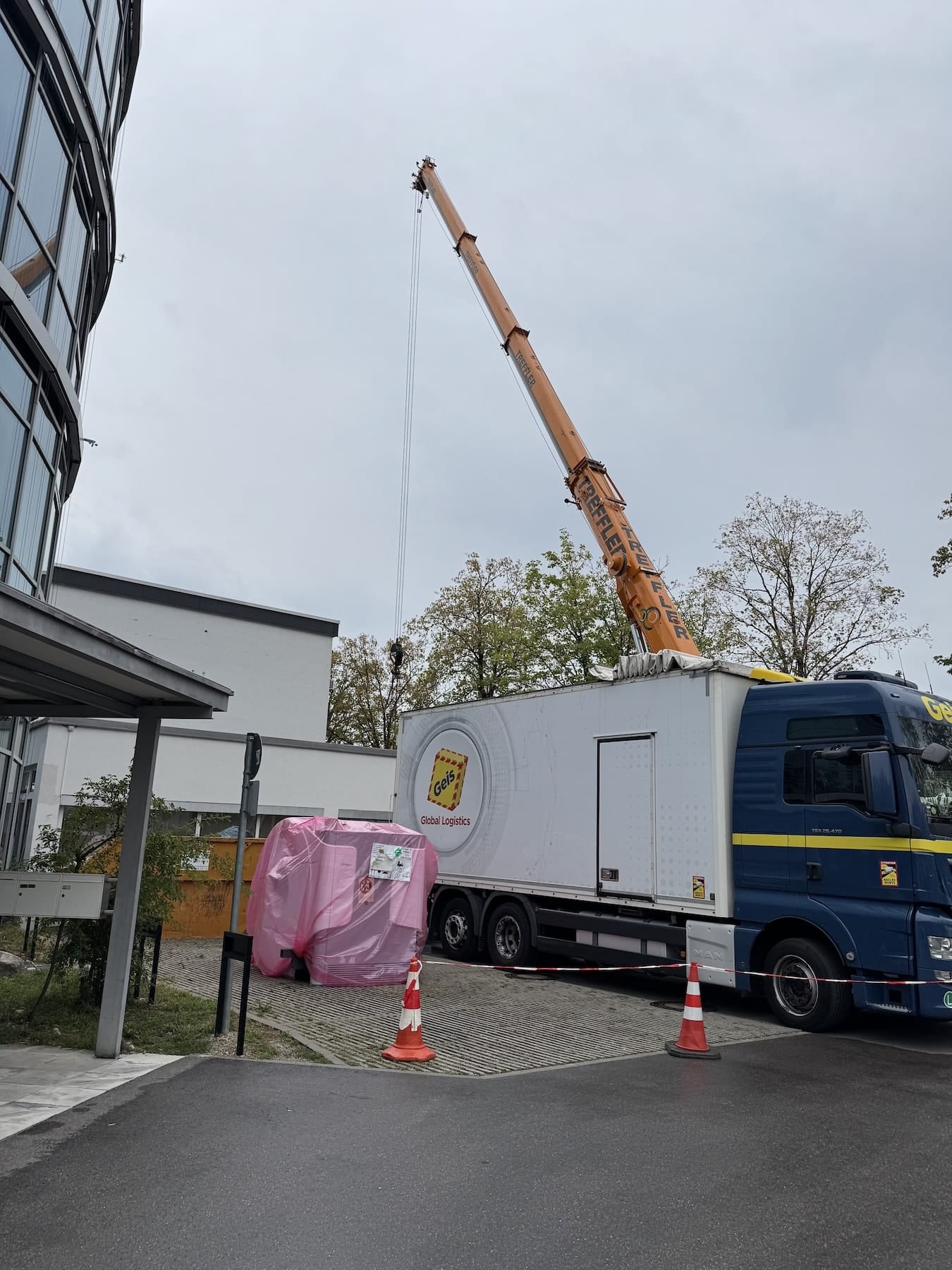 MRT-Anlieferung in Ottobrunn In rosa Schutzfolie verpackter MRT-Magnet neben einem Sattelzug vor dem Praxisgebäude in Ottobrunn
